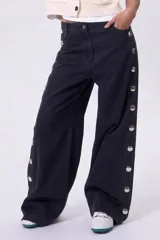 Pantalón de jean negro de corte wide leg, con detalle de botones metálicos plateados a lo largo de los laterales de ambas piernas.