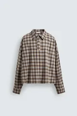 Camisa polo de corte boxy fit confeccionada en tejido de algodón con elasticidad, presenta un estampado de cuadros en tonos marrones, azules y blancos. Cuenta con cuello solapa, cierre frontal de botones, manga larga con puños abotonados y bajo con aberturas laterales.