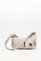 Cartera tipo shoulder color beige, de diseño utilitario y formato curvo. Confeccionada en material liviano y resistente, cuenta con correa regulable para llevar al hombro o cruzada. Presenta múltiples bolsillos con cierre en el frente y laterales, y un parche con el logo "Piece & Cake" en el frente.