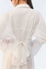 Camisa oversize negra de algodón liviano con textura calada, cuello camisero, botones frontales, mangas largas voluminosas y tiras para anudar en la espalda.