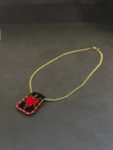 Collar con cadena dorada y dije rectangular de tela negra bordado a mano con un corazón rojo y detalles en hilo dorado, bordeado con mostacillas rojas.