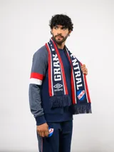 Bufanda tejida del Club Nacional de Football, con diseño tricolor en blanco, azul y rojo. Presenta inscripciones con letras grandes y el logo de Umbro en los extremos, finalizando con flecos decorativos.
