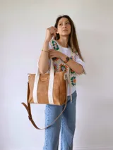 Cartera tipo tote de cuero color camel con detalles de bandas verticales en color crema. Cuenta con doble asa de mano, correa larga ajustable y extraíble, y cierre superior. Incluye etiqueta de cuero con logo grabado en el frente.
