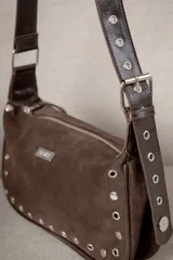 Cartera de hombro confeccionada en gamuza color bordó, con diseño estructurado y cierre superior. Presenta una correa ajustable de cuero con ojales metálicos y tachas decorativas a lo largo de la base y los laterales. Incluye una placa metálica con logo en el frente.