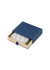 Billetera plegable de cuero color marrón claro, con costuras blancas visibles en los bordes. Presenta un pequeño logo grabado en la parte inferior derecha y una abertura rectangular cosida en la parte superior central. En el lateral derecho se aprecian detalles de tela con los colores de la bandera francesa (azul, blanco y rojo).