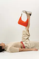 Cartera tipo shoulder bag tejida a mano con punto crochet en raffia color naranja. Tiene asa de cuero marrón y cierre superior con cremallera.