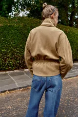 Chaqueta de gamuza sintética color marrón, con cuello subido, cierre frontal cruzado con botones y mangas largas anchas. Cuenta con bolsillos delanteros y cinturón ajustable del mismo tejido.