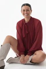 Blusa plisada de corte amplio y fluido, color bordó, con cuello alto y lazo decorativo. Presenta mangas largas con volumen y puños ajustados.