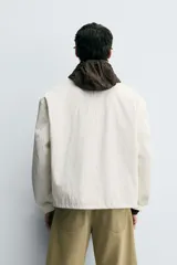 Cazadora de hombre, corte relaxed fit, color blanco hueso o crema, con acabado brillante. Presenta cuello solapa, manga larga, dos bolsillos de plastrón con solapa en la cadera y cierre frontal con botones a presión ocultos por una solapa.