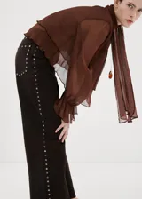 Pantalón de jean color marrón oscuro, de corte recto y tiro medio. Presenta una hilera de tachas metálicas plateadas a lo largo de la costura lateral externa.