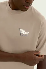 Remera beige de manga corta con cuello redondo, confeccionada en tejido de algodón. Presenta un pequeño estampado en el pecho con diseño de una bandera y el texto "H R NG est. 1880".