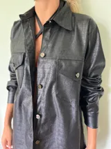 Camisa negra de cuero sintético con textura de cocodrilo, corte recto, cuello clásico, cierre frontal con botones metálicos y bolsillos con solapa en el pecho.
