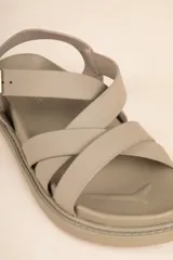 Sandalia baja de tiras cruzadas color camel, con hebilla metálica ajustable en el tobillo y suela negra gruesa.