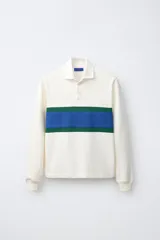 Remera polo de manga larga de corte regular fit, confeccionada en algodón color blanco crudo. Presenta cuello solapa blanco, cierre frontal con botonadura oculta y una franja horizontal ancha en el pecho en color azul y verde.
