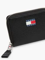 Billetera negra de cuero sintético con cierre de cremallera y logo de Tommy Jeans en el frente. Incluye compartimentos internos para tarjetas, billetes y monedas.