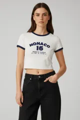 Remera de corte crop, color blanco con detalles en azul marino en cuello y mangas. Presenta un estampado frontal con la inscripción 'MONACO 16' y coordenadas geográficas.
