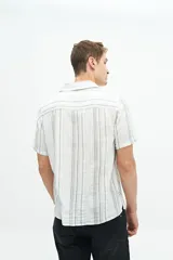 Camisa de manga corta con cuello cubano, confeccionada en una mezcla de lino y algodón con textura. Presenta un diseño a rayas verticales en tonos beige y celeste sobre fondo blanco, con cierre frontal de botones y logo bordado en el pecho.