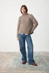 Sweater de punto acanalado en color gris topo, con cuello alto y mangas largas raglán. Presenta un diseño de corte holgado y una pequeña etiqueta decorativa en el lateral.