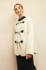 Abrigo gamulán de corderito color crudo, con alamares de cuero sintético negro y cierres metálicos.