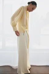 Pantalón wide leg de tiro medio confeccionado en hilatura de algodón, color beige, con trabillas en la cintura, bolsillos laterales ocultos y detalle de pliegues en el delantero. Cierre frontal con cremallera, botón y ganchos metálicos.