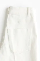 Jean de denim de algodón color blanco, de tiro alto y piernas amplias con corte holgado. Cuenta con pliegues frontales y traseros, cierre frontal con botón y bolsillos traseros de parche.