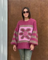 Buzo de tejido de punto con textura peluda, de corte holgado y mangas amplias. Presenta un diseño geométrico con una flor grande estampada en el centro, combinando tonos fucsia, rosa y marrón en un patrón de bloques y rayas.