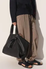 Bolso tipo shopper de cuero negro con textura granulada. Presenta doble asa de mano, cierre magnético y un detalle decorativo de nudo con argolla metálica en el frente.