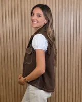 Chaleco de denim color marrón, de corte holgado, con cuello camisero, cierre frontal de botones metálicos y bolsillos appliqués con solapa en el pecho.