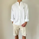 Camisa de lino blanca con cuello mao y cierre frontal de botones. Presenta un pequeño bordado oscuro en el pecho y mangas largas con puños doblados.