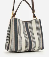 Cartera bucket pequeña de Satinato con diseño de rayas verticales en tonos blanco, azul marino y beige, confeccionada en tejido estructurado. Cuenta con asa de mano fija, asa larga desmontable de cuero sintético marrón, detalles de herrajes metálicos dorados, argolla metálica colgante en el frente y cierre superior.