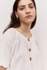 Blusa blanca de manga corta, con escote redondo y cierre frontal con tres botones. Presenta pliegues frontales y diseño liviano.