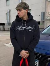 Campera con capucha negra, con cierre frontal completo y bolsillos estilo canguro. Presenta un estampado gráfico en blanco con letras cursivas en el pecho y diseño holgado.