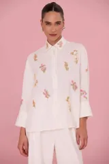 Camisa de lino de mujer color marfil, de corte recto y holgado, con cuello camisero y mangas largas terminadas en puños anchos. Presenta detalles de bordados florales dispersos en el frente y el cuello.