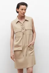 Vestido corto color beige, sin mangas, con cuello camisero, cierre frontal con cordones cruzados y múltiples bolsillos con cierre.