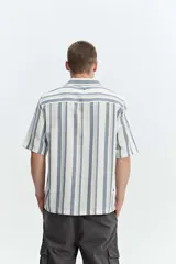 Camisa de manga corta oversize en tejido rústico, con diseño de rayas verticales celestes y blancas, cuello clásico y cierre frontal de botones.