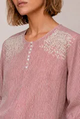 Blusa a rayas verticales en tonos rosados y blancos, con cuello redondo y cierre frontal parcial con botones. Presenta delicados detalles bordados en los hombros y mangas tres cuartos abullonadas con puños elásticos.
