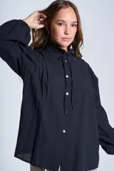 Camisa de color azul con cuello alto decorado con volados y lazo de ajuste. Presenta tablas decorativas en los hombros, cierre frontal con botones de nácar y mangas largas con puños elásticos que generan un efecto abullonado.