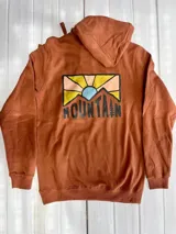 Hoodie color terracota/óxido con capucha y estampado gráfico en la espalda que representa un paisaje montañoso con un sol naciente o poniente y la palabra "MOUNTAIN" en la parte inferior.