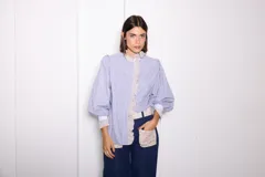 Camisa de manga larga con diseño combinado en tres telas, presentando un patrón de rayas verticales en azul y blanco. Cuenta con cuello alto, volados frontales y puños en contraste. El diseño es holgado con mangas abullonadas.