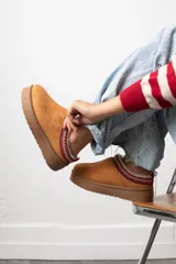 Zueco de gamuza color tostado con interior de corderito y detalle de cinta bordada en el borde superior. Cuenta con suela de plataforma plana de goma.