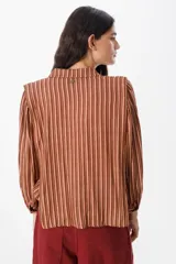 Camisa de manga larga con diseño de rayas verticales en tonos verde, azul y marrón. Presenta cuello clásico, cierre frontal con botones y mangas abullonadas con puños ajustados.