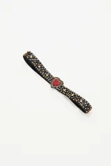 Cinturón elástico negro con hebilla en forma de corazón recubierta de strass rojos. La correa presenta aplicaciones de tachas metálicas y cristales en forma de estrellas y círculos.