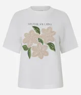 Remera de algodón color blanco, con cuello redondo y manga corta. Presenta un estampado con la frase 'Soy flor. Soy latina' y flores bordadas en relieve con textura tejida.