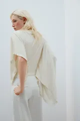 Camiseta tipo capa oversize de color blanco, confeccionada en hilatura de algodón 100%. Presenta cuello redondo, manga corta y un diseño de bajo asimétrico con caída pronunciada en uno de los laterales.