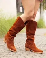 Bota estilo texana de caña alta, confeccionada en cuero gamuzado color marrón óxido. Presenta un diseño de corte en V en la parte superior de la caña, costuras decorativas y un taco bajo de madera.