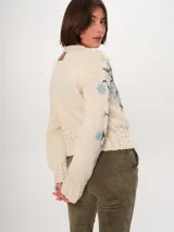 Cárdigan tejido a mano color crudo, con diseño de trenzas y flores bordadas en azul y marrón. Tiene cuello redondo y cierre frontal con botones de madera.