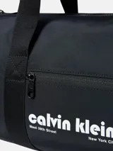 Bolso deportivo tipo duffle bag de color negro, confeccionado en poliéster. Presenta asas de mano y una correa ajustable para llevar al hombro. En el centro tiene un bolsillo frontal con cierre donde se lee el logo de Calvin Klein en blanco, incluyendo las direcciones "West 39th Street" y "New York City".