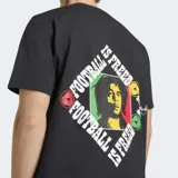 Remera negra de algodón con cuello redondo y mangas cortas. Presenta el logo del Trifolio de Adidas en blanco en el pecho derecho y un parche circular con manos entrelazadas en colores rasta en el pecho izquierdo. Incluye un pequeño parche rectangular en la parte inferior derecha.