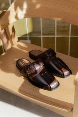 Sandalias tipo mule de cuero marrón con diseño cerrado en la puntera y aberturas laterales, con detalle de hebilla lateral y costuras a la vista.