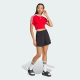 Remera corta roja ajustada al cuerpo, de manga corta, con cuello redondo blanco y las icónicas tres líneas blancas de Adidas en las mangas. Cuenta con el logo del trifolio bordado en el pecho.